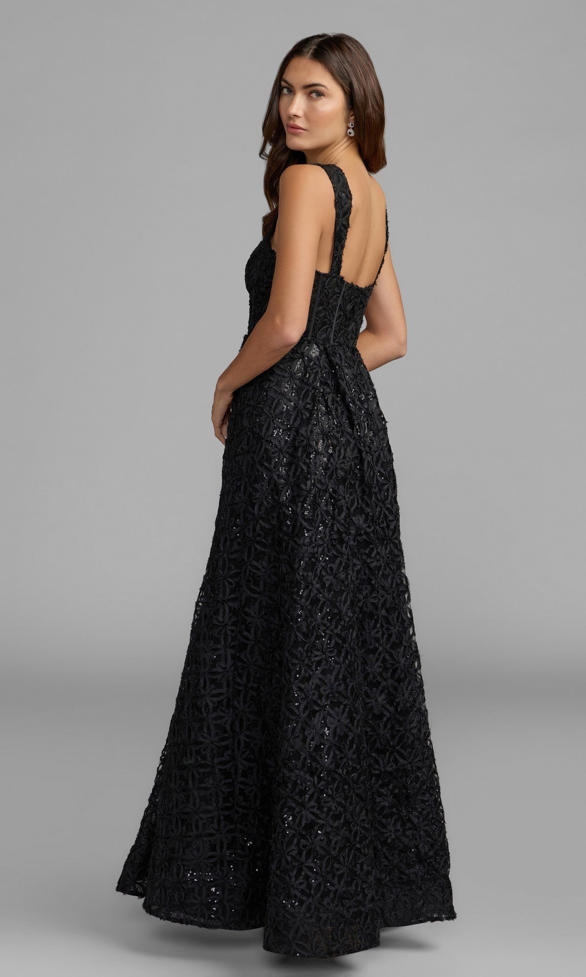 A woman in the Long Formal Dress: Mac Duggal 56286—a sleeveless, floor-length black gown featuring textured geometric patterns—stands facing away, glancing over her shoulder.